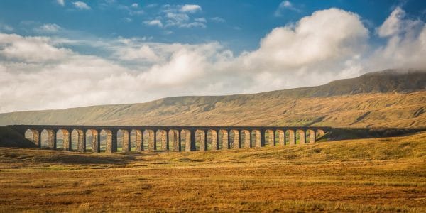 Panoramic photograph of Ribblehead Viaduct in the Yorkshire Dales, with Whernside in the background, golden hillsides and a blue sky with white clouds.