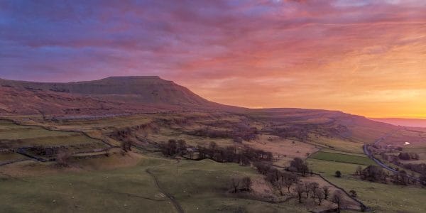 Aerial panoramic photograph of Ingleborough in the Yorkshire Dales at sunset, with pink, purple and orange skies above the mountain and surrounding fields.