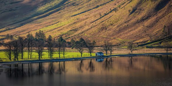 Panoramic photograph of Buttermere in the Lake District, with autumn sunshine illuminating the fells, lakeside trees and reflections in the water.